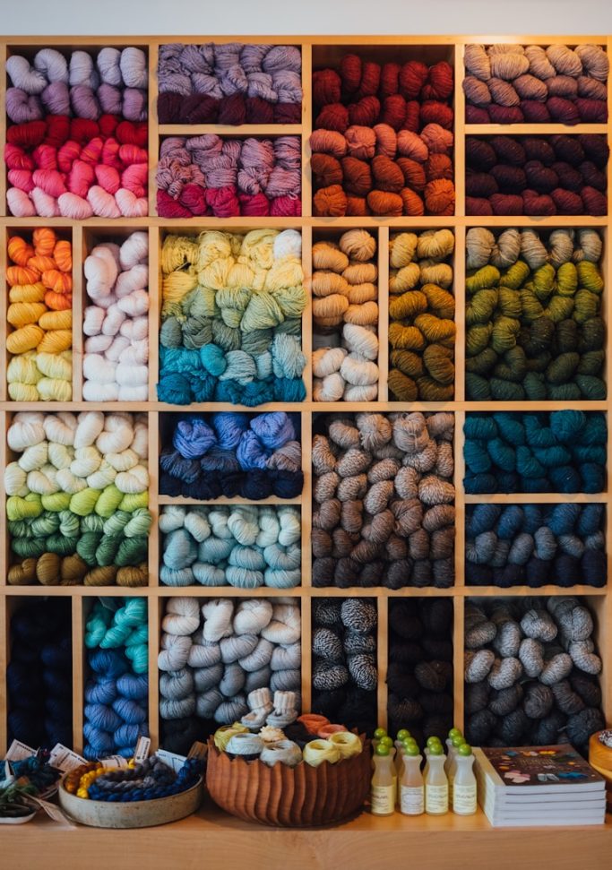 Bunte Wolle in Regalen, geordnet nach Farben von Rot bis Blau und einer Auswahl an Zubehör.